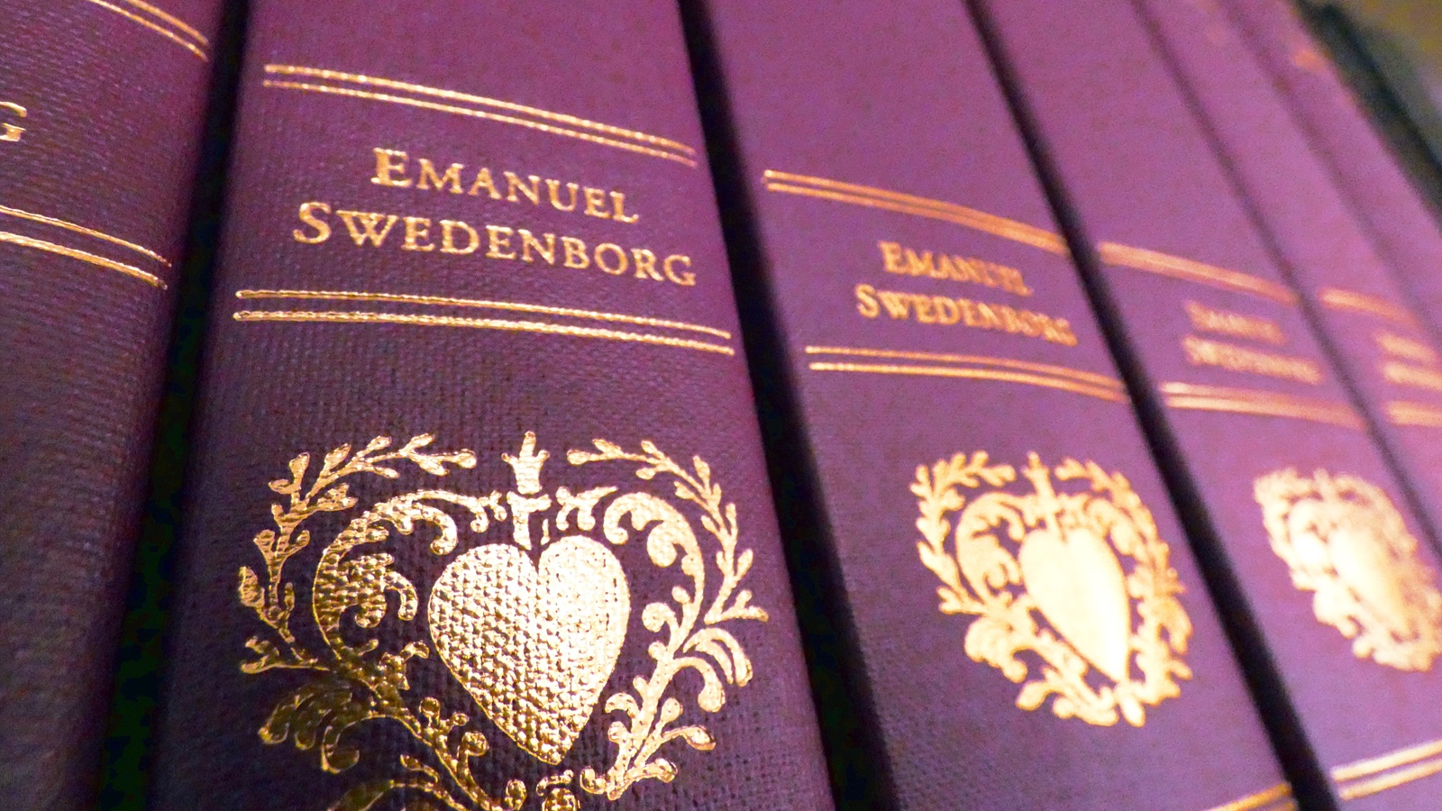 A close-up photograph of a series of purple hardcover New Century Edition books. Shiny gold lettering reads "Emanuel Swedenborg" and displays a stylized heart ornament.