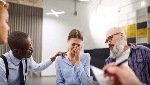 A group of people sit in a white room. A woman appears to be crying in the center of the image, she is comforted by a man with glasses on the left, and a man with a large white beard on the right.