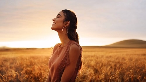 A woman with long brown hair and large hoop earrings stands in a golden field, looking contentedly off into the sunset.