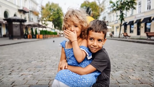 A young girl child wearing a blue eyelet dress sits on the lap of a young boy child wearing a dark grey t-shirt.